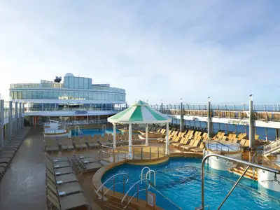 Das Pooldeck der Norwegian Jewel bei Tageslicht. Blaue Pools, Whirlpools und viele Liegestühle. Moderne Glasfassaden des Schiffes, das weite Meer und blauer Himmel.