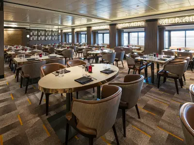 Das ChinChin Spezialitätenrestaurant an Bord der Norwegian Jewel zeigt eine Reihe von leeren, stilvoll gedeckten Tischen und Stühlen in einem modern eingerichteten Raum. Große Fenster mit Jalousien spenden Tageslicht.