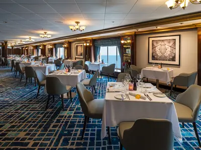 Elegantes Restaurant an Bord der Norwegian Jewel. Weiße Tische, blauer Teppich. Meerblick durch große Fenster. Gediegene, einladende Atmosphäre. Geschirr und Gläser bereit.