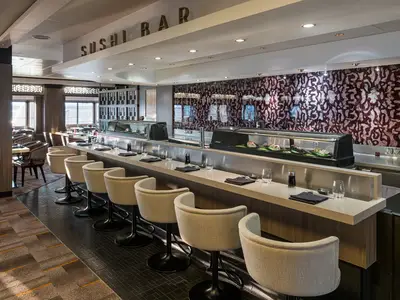 Elegante Sushi-Bar an Bord der Norwegian Jewel. Eine lange Theke mit modernen Barhockern, Gedecken und beleuchteter Vitrine mit frischem Sushi. Im Hintergrund weitere Tische des Restaurants.