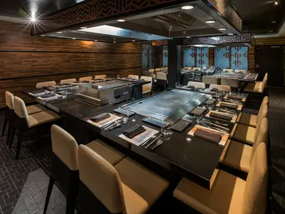 Blick in das Teppanyaki Restaurant der Norwegian Jewel. Schwarze Granittische mit Kochplatten und beigefarbenen Stühlen sind gedeckt. Moderne Holzwände.