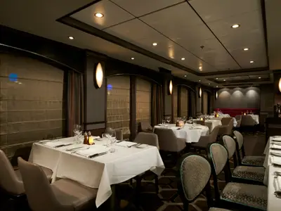 Ein stilvolles Spezialitätenrestaurant, Cagney's Steakhouse, an Bord der Norwegian Jewel mit mehreren gedeckten Tischen, beigen Stühlen und indirekter Beleuchtung. Dunkle Holzelemente und helle Wandleuchten prägen das Ambiente.