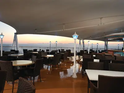 Aussenrestaurant der Norwegian Jewel am Abend. Der überdachte Bereich zeigt Tische, Stühle und Laternen. Im Hintergrund sind Meer und entfernte Berge beim Sonnenuntergang zu sehen.