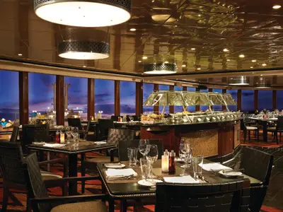 Elegantes Spezialitätenrestaurant der Norwegian Jewel. Gedeckte Tische, Buffet und große Fenster mit Blick auf das Meer und einen beleuchteten Hafen bei Sonnenuntergang.