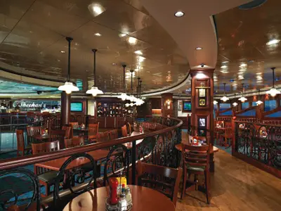 Das O'Sheehan's Restaurant an Bord der Norwegian Jewel zeigt eine gemuetliche Einrichtung mit dunklem Holz, Tischen, Stühlen und einer Bar im Hintergrund. Der Bereich ist hell erleuchtet mit vielen Pendelleuchten und Downlights.