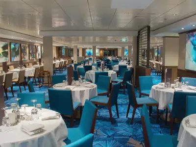 Geräumiges Restaurant Azura an Bord der Norwegian Jewel. Festlich gedeckte Tische mit blauen Stühlen auf blauem Teppich. Moderne Kunst an den Wänden. Helle Atmosphäre.