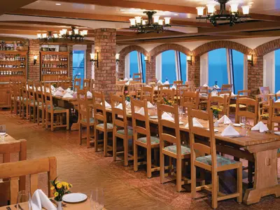 Ein rustikales Restaurant an Bord der Norwegian Jewel mit langen Holztischen und Stühlen. Backsteinsäulen und Bögen prägen den Raum. Große Fenster bieten einen weiten Blick auf das blaue Meer.