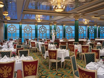 Blick in den luxuriösen Speisesaal 'Tsar's Palace' an Bord der Norwegian Jewel. Zahlreiche Tische sind festlich gedeckt. Die Stühle sind in Rot und Grün gehalten und mit goldenen Doppeladler-Motiven verziert. Große runde Fenster mit Ornamenten bieten einen weiten Blick auf das blaue Meer. Goldene Kronleuchter und grüne Marmorsäulen prägen die opulente Einrichtung.