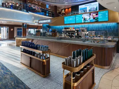 Ein moderner Starbucks an Bord der Norwegian Jewel mit einer langen Theke, digitaler Speisekarte und blauen Fliesen. Im Vordergrund stehen Verkaufsregale mit Tassen und Bechern.