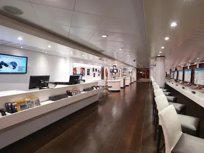 Eine lange, moderne Galerie mit Verkaufsregalen und Bildschirmen auf der linken Seite. Rechts sind mehrere Computer-Terminals mit Stühlen für Gäste an Bord der Norwegian Jewel zu sehen.