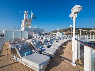 Ein sonniges Deck an Bord der Norwegian Jewel mit Reihen von grauen Liegestühlen und kleinen Tischen, einige mit Getränken und Handtüchern. Im Hintergrund sind Berge zu sehen.