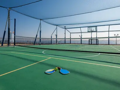 Grüner Sportplatz an Bord der Norwegian Jewel mit Pickleball-Schlägern und Ball. Netz, Basketballkorb und Tor. Hohe Schutznetze umgeben den Bereich mit Meerblick unter blauem Himmel.