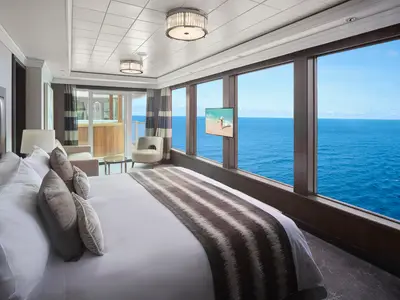 Luxuriöse Suite der Norwegian Jewel. Helles Schlafzimmer mit Doppelbett, Sitzbereich und grossen Fenstern zum blauen Meer. Links ein privater Balkon mit Whirlpool.