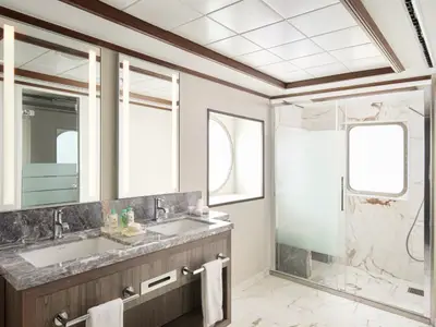 Helles, luxuriöses Badezimmer an Bord der Norwegian Jewel. Es verfügt über ein Doppelwaschbecken, eine geräumige Dusche mit Glaswand und zwei Bullaugen für Tageslicht.