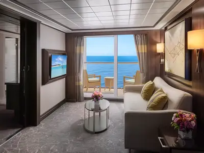 Eine luxuriöse Suite mit Wohnbereich, Sofa und Balkon mit Meerblick auf der Norwegian Jewel. Links ein Fernseher, rechts Wandleuchten und Kunst. Ein Couchtisch mit Blumen steht im Vordergrund.