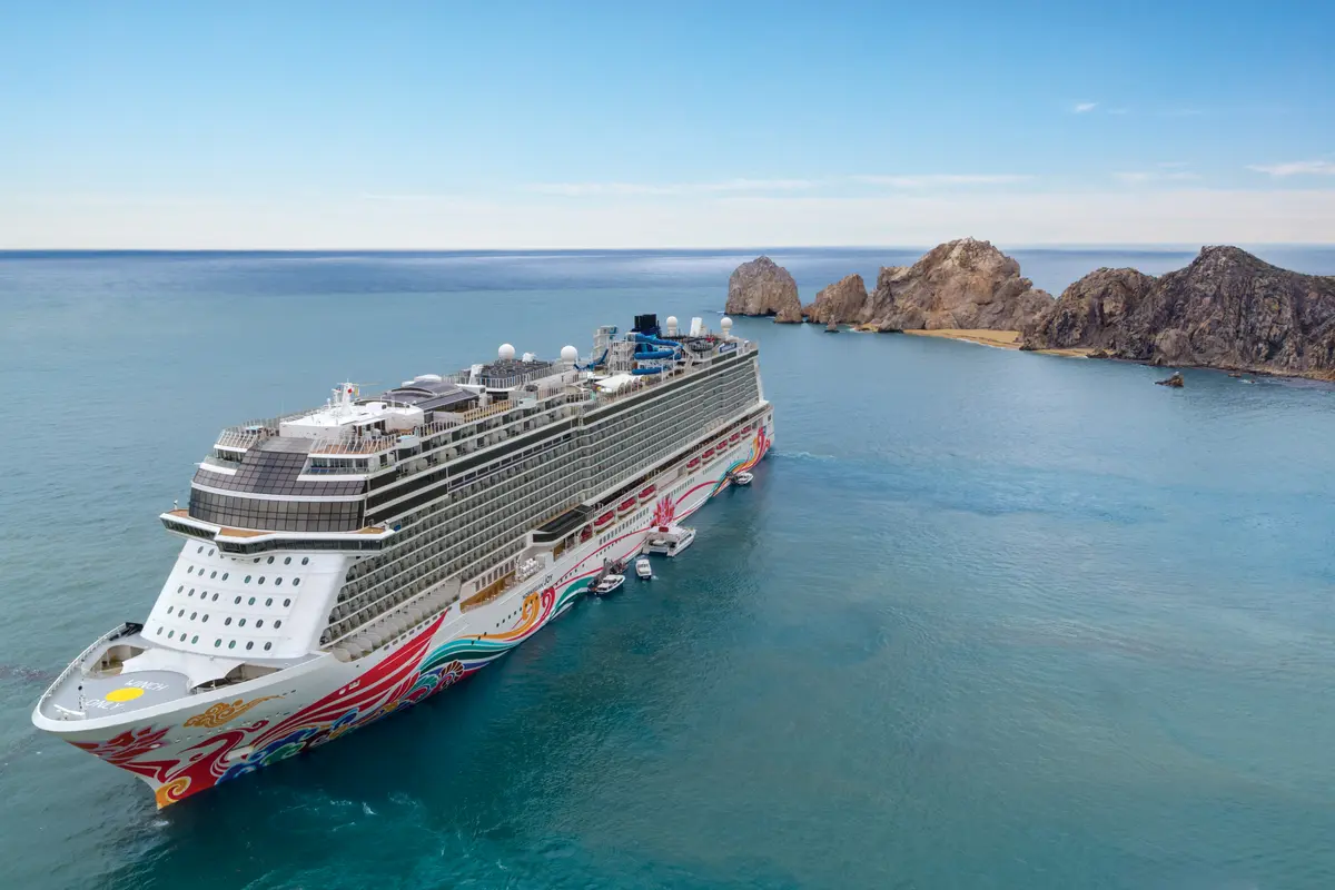 Luftaufnahme der Norwegian Joy auf dem Meer. Das Kreuzfahrtschiff mit bunter Bugbemalung fährt an den Felsformationen von Cabo San Lucas vorbei. Blauer Himmel und ruhiges Wasser.