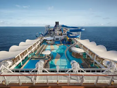 Vogelperspektive auf das lebhafte Pooldeck der Norwegian Joy mit blauen Wasserrutschen, Pools und einem farbenfrohen Wasserpark. Zahlreiche Liegestühle laden zur Entspannung unter dem weiten Himmel ein. Ein idealer Ort für Familien an Bord der Norwegian Joy.