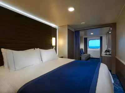 Eine helle, moderne Aussenkabine an Bord der Norwegian Joy mit Doppelbett, blauer Tagesdecke und einem runden Fenster, das den Blick auf das blaue Meer freigibt.