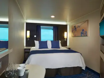 Aussenkabine der Norwegian Joy: Doppelbett, blaue Kissen, Meerblick durch Fenster. Moderne Einrichtung, Kunst, indirekte Beleuchtung. Gemütlicher Raum.