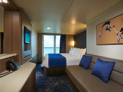 Blick in eine moderne Balkonkabine der Norwegian Joy mit Doppelbett, blauer Tagesdecke, grauem Sofa, Flachbildfernseher und Fenster mit Meerblick.