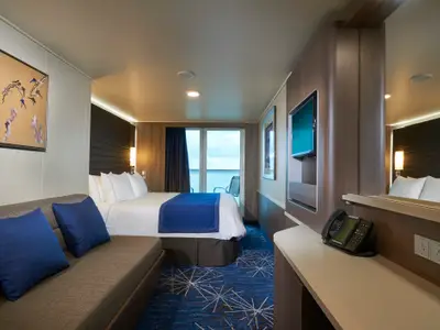 Eine helle, moderne Balkonkabine an Bord der Norwegian Joy mit einem grossen Bett, blauer Tagesdecke, einem grauen Sofa mit blauen Kissen und einem Balkon mit Meerblick.