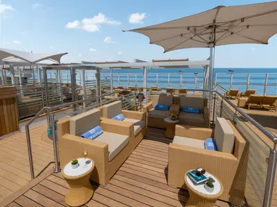 Outdoor-Lounge auf der Norwegian Joy. Rattan-Sitzgruppen mit NCL-Handtüchern, Sekt, Obst. Panoramablick auf das blaue Meer unter weitem Himmel.
