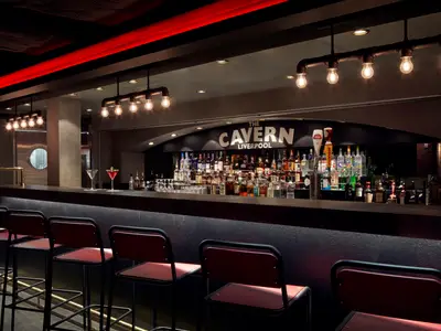 Eine stilvolle Bar namens 'The Cavern Liverpool' an Bord der Norwegian Joy. Der Barbereich ist reich mit Spirituosen bestueckt, davor stehen rote Barhocker und moderne Industrieleuchten spenden warmes Licht.