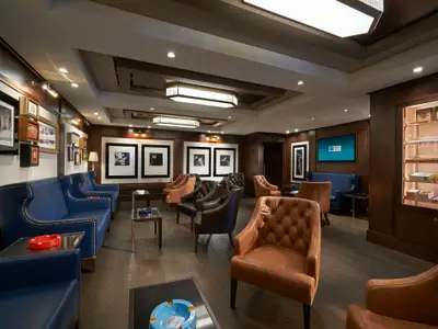 Blick in die elegante Zigarren-Lounge an Bord der Norwegian Joy. Der Raum ist mit bequemen blauen Sofas, braunen Ledersesseln, dunklen Holzwänden und gerahmter Kunst ausgestattet. Eine beleuchtete Vitrine zeigt Zigarren.
