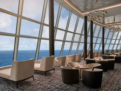 Blick in die moderne Observation Lounge an Bord der Norwegian Joy. Der Raum ist mit eleganten Sesseln und Tischen ausgestattet. Große, schräge Fenster bieten einen weiten Blick auf das offene Meer und den bewölkten Himmel.
