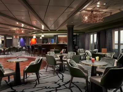 Stilvolle Bar-Lounge an Bord der Norwegian Joy mit bequemen Sesseln, runden Tischen und einer Bar. Große Fenster geben den Blick auf das Meer frei. Mehrere bunte Cocktails sind zu sehen.