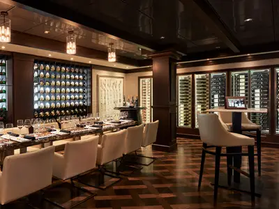 Die elegante Weinbar 'The Cellars' an Bord der Norwegian Joy. Ein langer Holztisch mit vielen Weingläsern ist mittig platziert, umgeben von Weinregalen an den Wänden. Rechts stehen Stehtische mit Barhockern.