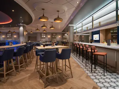Blick auf "The Local Bar & Grill" an Bord der Norwegian Joy. Dunkelblaue Hochstühle, Holztische, eine Bar mit orangefarbenen Hockern und gemustertem Boden.