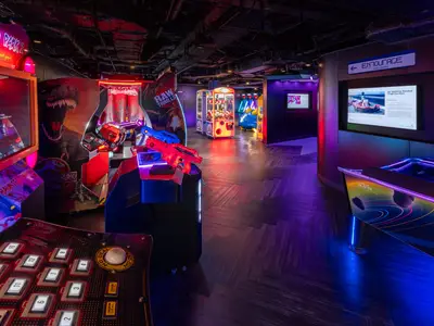 Eine lebhafte Spielhalle an Bord der Norwegian Joy. Im Vordergrund sind beleuchtete Arcade-Automaten mit roten und blauen Gewehren zu sehen. Im Hintergrund stehen weitere Spielautomaten und eine Beschilderung zum 'ENTOURAGE TEEN LOUNGE'.