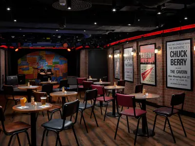 Innenansicht des Cavern Club an Bord der Norwegian Joy. Bühne mit Schlagzeug, bunter Wandkunst. Tische, Stühle, Getränke sind im Raum. Ziegelwände zeigen Musikplakate und haben gedämpftes Licht.