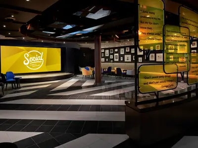 Ein moderner Comedy- und Nachtclub, der Social, an Bord der Norwegian Joy. Eine Bühne mit grosser gelber LED-Wand, davor blaue Stühle. Im Hintergrund Lounge-Bereiche und eine Wand mit Bildern.