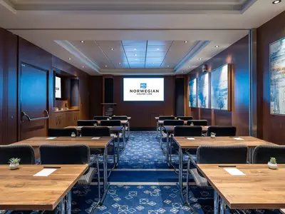 Ein leerer Konferenzraum an Bord der Norwegian Joy mit holzgetäfelten Wänden, blauem Teppich, dunklen Stühlen und Holztischen. An der Stirnseite ein großer Bildschirm mit dem Logo von Norwegian Cruise Line.