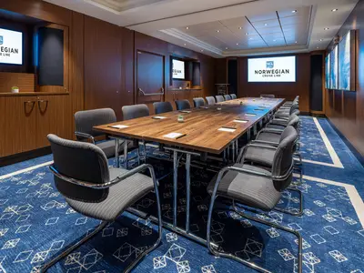 Moderner Konferenzraum an Bord der Norwegian Joy. Langer Holztisch, graue Stühle, großer Bildschirm mit NCL-Logo. Holzvertäfelte Wände, blauer Teppich. Professionelles Ambiente.