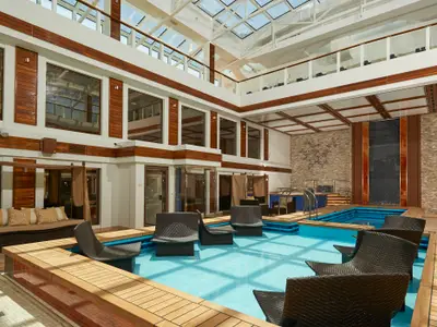 Ein luxuriöser Innenpool-Bereich an Bord der Norwegian Joy, umgeben von Holzelementen, großen Fenstern und bequemen Lounge-Stühlen im Wasser. Die Decke ist teilweise verglast und lässt viel Tageslicht herein.