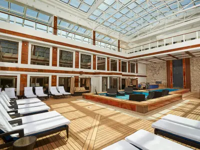 Luxuriöser Haven Courtyard Poolbereich an Bord der Norwegian Joy. Ein Glasdach überspannt das Holzdeck mit Pool, Lounge-Möbeln und Sonnenliegen.
