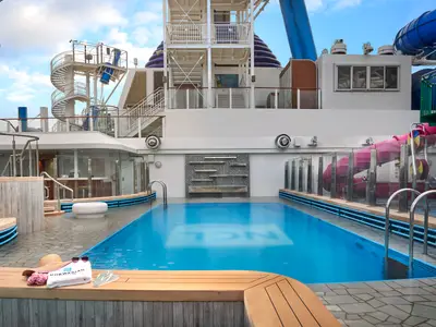 Hauptpoolbereich der Norwegian Joy: Blauer Pool, umgeben von Decks. Wasserrutschen und obere Decks sichtbar. Eine Holzbank mit Handtuch und Hut im Vordergrund.
