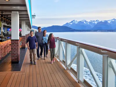 Eine Familie läuft lachend auf der Promenade der Norwegian Joy. Im Vordergrund eine Bar mit Ziegeln, im Hintergrund Berge und das Meer bei Tageslicht.