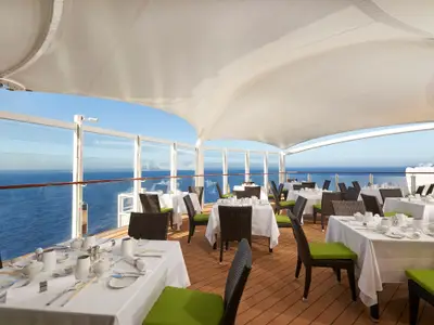 Das Aussenrestaurant an Bord der Norwegian Joy: stilvolle Tische unter einem Sonnensegel mit Panoramablick auf den blauen Ozean und den Horizont.
