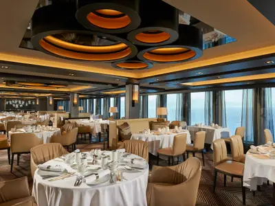 Blick in das elegante Restaurant an Bord der Norwegian Joy. Es gibt viele gedeckte Tische, bequeme Stühle und eine Fensterfront mit Meerblick. Warme Deckenbeleuchtung.