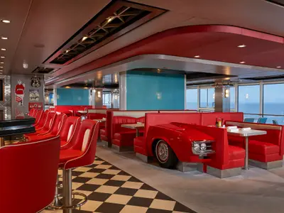 Das lebhafte 50er-Jahre American Diner an Bord der Norwegian Joy. Rote Barhocker und gepolsterte Sitzbänke, teilweise als Auto-Thema gestaltet, prägen den Raum. Der schwarz-weiß karierte Boden und große Fenster mit Meerblick ergänzen das Retro-Ambiente.