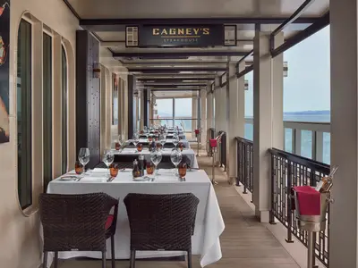 Blick in das elegante Cagney's Steakhouse an Bord der Norwegian Joy. Der überdachte Restaurantbereich bietet viele stilvoll gedeckte Tische mit weitem Meerblick auf eine Küstenlinie.