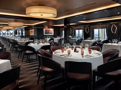 Ein elegantes Restaurant an Bord der Norwegian Joy mit runden Tischen, weissen Tischdecken und Lederstuehlen. Zahlreiche Weinglaeser und Geschirr sind eingedeckt. Grossflaechige Fenster geben den Blick auf das Meer frei.
