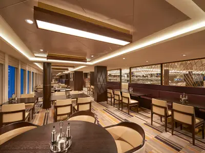 Ein helles und modernes Buffet-Restaurant namens Garden Cafe an Bord der Norwegian Joy. Der Raum ist mit dunklen Holztischen, beigen Stühlen und langen Polsterbänken ausgestattet. Entlang der linken Seite erstrecken sich große Fenster. Im Hintergrund ist der Buffetbereich mit Speisen hinter einer dekorativen Trennwand zu sehen. Das Restaurant ist leer.