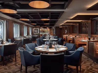 Blick in ein stilvolles Spezialitätenrestaurant an Bord der Norwegian Joy. Gedeckte Tische, blaue Ledersessel, braune Sitzbänke und große Fenster säumen den Raum. Keine Personen sichtbar.