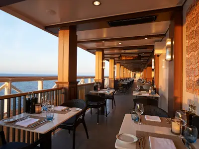 Ein stimmungsvolles Foto des La Cucina Spezialitätenrestaurants an Bord der Norwegian Joy mit Tischen auf dem Außendeck bei Sonnenuntergang. Das Meer ist im Hintergrund sichtbar.