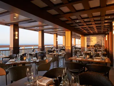 Elegantes Restaurant an Bord der Norwegian Joy mit stilvollen Tischen, warmer Beleuchtung und kunstvoller Fliesenwand. Blick auf das Meer bei Sonnenuntergang.